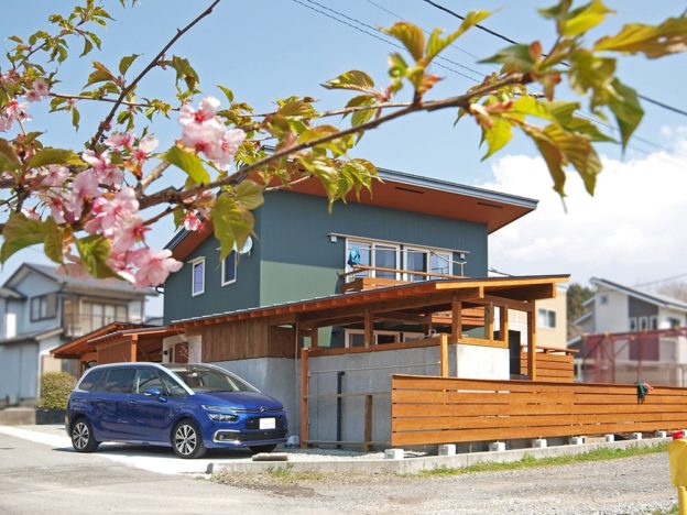 河津桜と緑のガルバリウムの外壁がよく馴染む。DIYによるアウトドアキッチンや物置の片流れが母屋の屋根と折り重なって美しい。